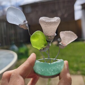 May include: A handcrafted glass flower sculpture with four sea glass blooms in white, pink, and green. The flowers are set in a small, turquoise sea glass pot. The sculpture is held in a person's hand.