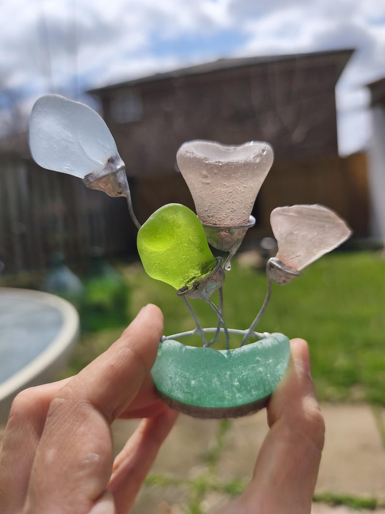 May include: A handcrafted glass flower sculpture with four sea glass blooms in white, pink, and green. The flowers are set in a small, turquoise sea glass pot. The sculpture is held in a person's hand.