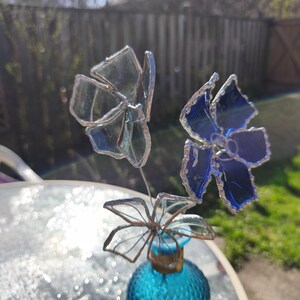 May include: A decorative glass flower arrangement. The arrangement features three flowers in shades of blue and clear glass. The flowers are attached to a small, textured, turquoise glass bottle. The arrangement is set outdoors.