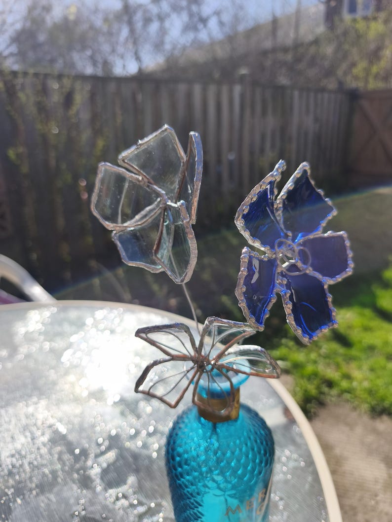 May include: A decorative glass flower arrangement. The arrangement features three flowers in shades of blue and clear glass. The flowers are attached to a small, textured, turquoise glass bottle. The arrangement is set outdoors.