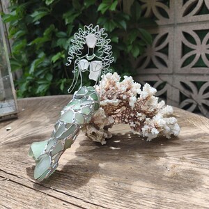 Puede incluir: Una figura decorativa de sirena hecha de alambre plateado y piezas de vidrio marino verde p&aacute;lido. La sirena presenta una cabeza y cabello esculpidos en alambre, un corpi&ntilde;o de vidrio marino y una cola hecha de fragmentos de vidrio verde superpuestos, apoyada contra un trozo de coral blanco.