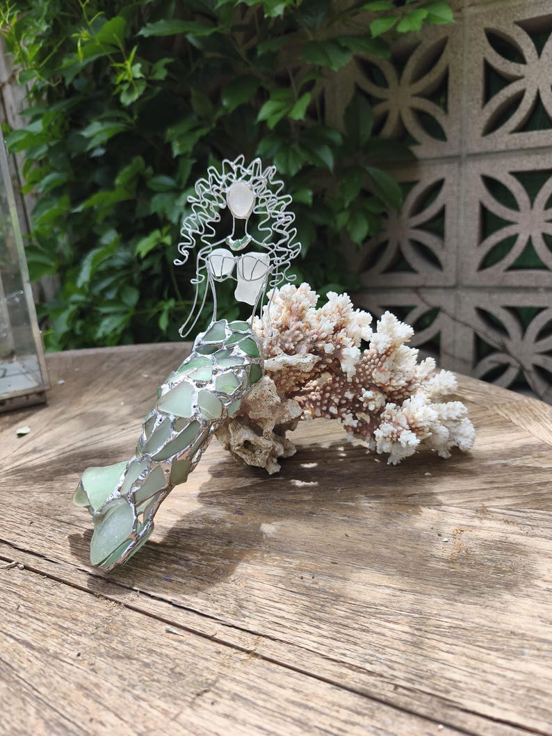 Puede incluir: Una figura decorativa de sirena hecha de alambre plateado y piezas de vidrio marino verde p&aacute;lido. La sirena presenta una cabeza y cabello esculpidos en alambre, un corpi&ntilde;o de vidrio marino y una cola hecha de fragmentos de vidrio verde superpuestos, apoyada contra un trozo de coral blanco.
