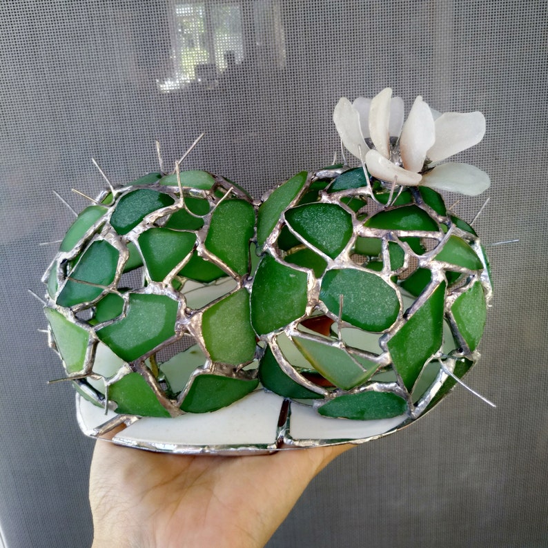 May include: A green glass cactus sculpture with silver accents and a white flower on top. The sculpture is made of many small pieces of glass that are joined together to create a three-dimensional design.