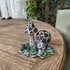 May include: A decorative mushroom sculpture with two mushroom caps. The caps are brown with a mosaic pattern, and the stems are silver. The base is green and silver, resembling moss. The sculpture is on a wooden table.