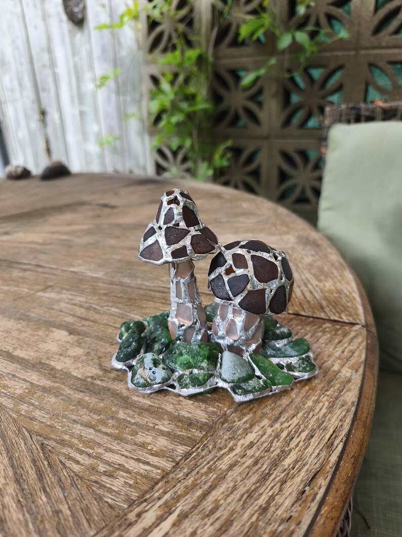 May include: A decorative mushroom sculpture with two mushroom caps. The caps are brown with a mosaic pattern, and the stems are silver. The base is green and silver, resembling moss. The sculpture is on a wooden table.