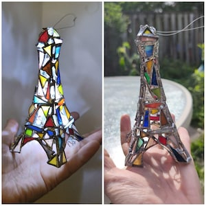 May include: A colourful stained glass Eiffel Tower ornament. The tower is made from pieces of red, yellow, blue, and green glass, held together by metal. A wire loop is attached for hanging, perfect for a window display.