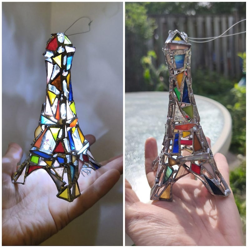 May include: A colourful stained glass Eiffel Tower ornament. The tower is made from pieces of red, yellow, blue, and green glass, held together by metal. A wire loop is attached for hanging, perfect for a window display.
