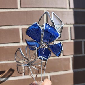 May include: A handcrafted stained glass flower sculpture with vibrant blue petals and clear accents. The flower is held by a wire stem, showcasing the intricate details of the glasswork. The sculpture is set against a brick wall background.