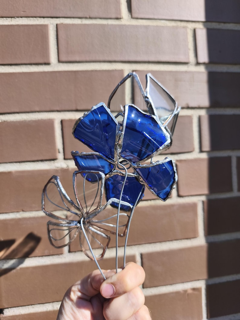 May include: A handcrafted stained glass flower sculpture with vibrant blue petals and clear accents. The flower is held by a wire stem, showcasing the intricate details of the glasswork. The sculpture is set against a brick wall background.