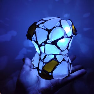 May include: A small, white, stained glass lamp with a blue light shining through it. The lamp is made of many small pieces of glass, some of which are yellow. The lamp is being held in a person's hand.