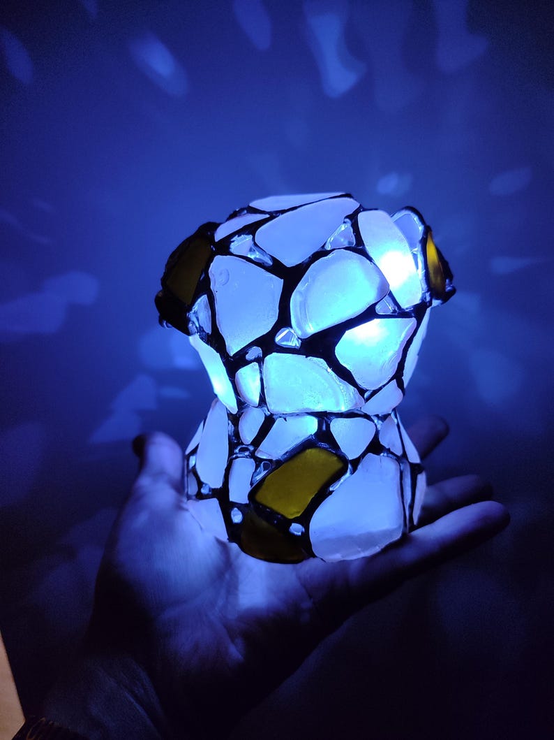May include: A small, white, stained glass lamp with a blue light shining through it. The lamp is made of many small pieces of glass, some of which are yellow. The lamp is being held in a person's hand.