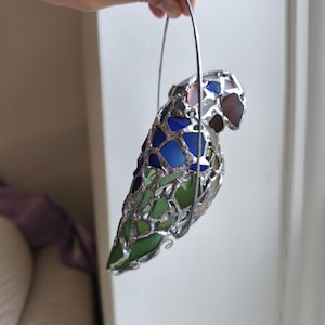 May include: A stained glass parrot sculpture with a silver metal frame. The parrot is made of blue, green, and brown glass pieces. It is held by a silver metal handle.