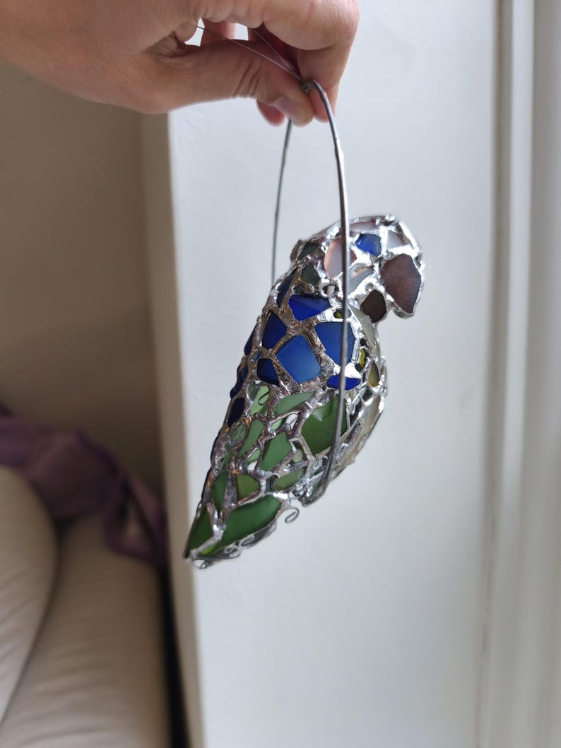 May include: A stained glass parrot sculpture with a silver metal frame. The parrot is made of blue, green, and brown glass pieces. It is held by a silver metal handle.