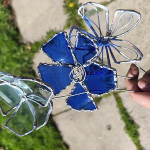 May include: A handcrafted stained glass flower sculpture with three flowers in blue and green hues. The petals are outlined in silver solder, and the sculpture is held by a wire stem. The background shows green grass and a concrete surface.