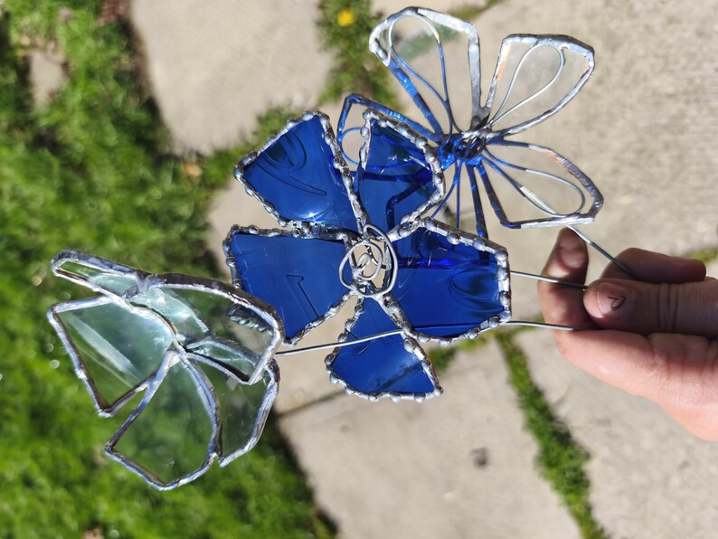 May include: A handcrafted stained glass flower sculpture with three flowers in blue and green hues. The petals are outlined in silver solder, and the sculpture is held by a wire stem. The background shows green grass and a concrete surface.