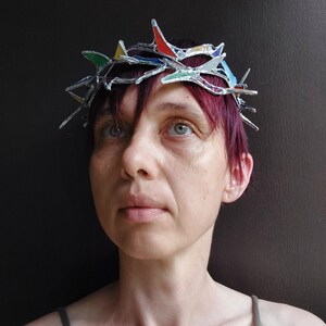 May include: A crown made of silver-toned metal and small, multi-colored stained glass shards in red, yellow, green, and blue. The structure features sharp, pointed protrusions arranged in a circular band that rests upon the head.
