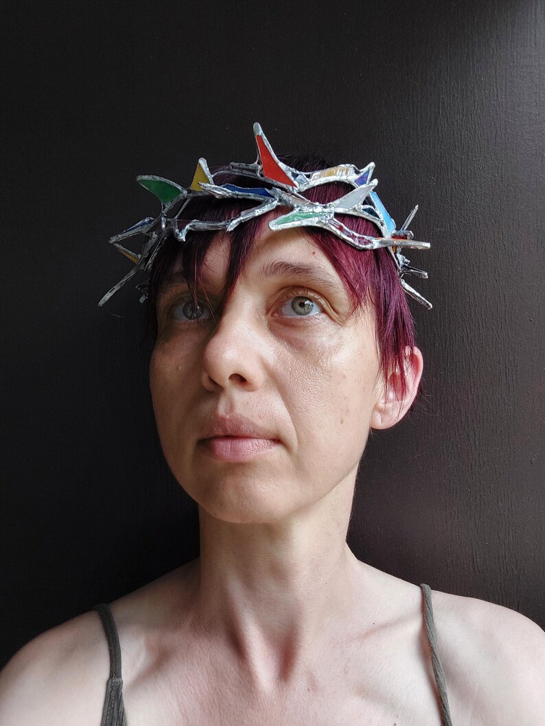 May include: A crown made of silver-toned metal and small, multi-colored stained glass shards in red, yellow, green, and blue. The structure features sharp, pointed protrusions arranged in a circular band that rests upon the head.