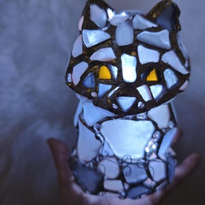 May include: A mosaic cat ornament with a glowing interior. The cat is made from white, grey, and blue glass pieces, with yellow eyes. The figurine is held in a hand, and the light from within illuminates the glass, creating a soft glow.