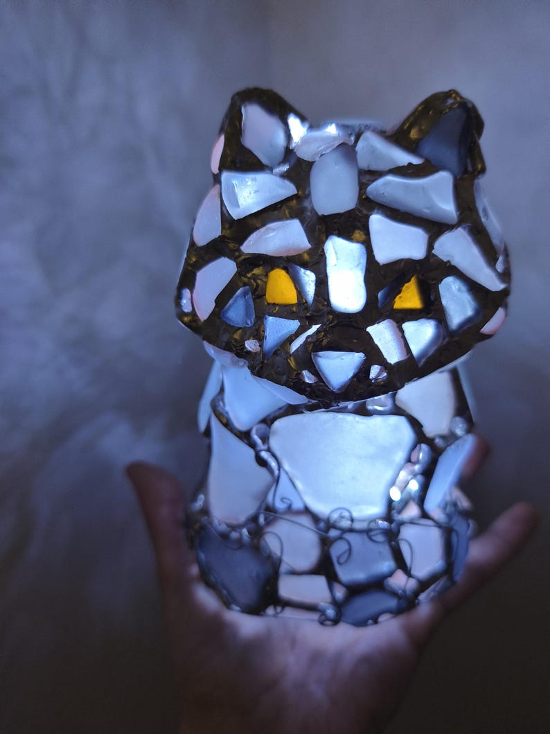 May include: A mosaic cat ornament with a glowing interior. The cat is made from white, grey, and blue glass pieces, with yellow eyes. The figurine is held in a hand, and the light from within illuminates the glass, creating a soft glow.