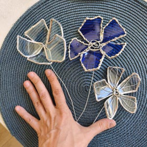 May include: Three stained glass flower sculptures. One is clear, one is blue, and one is clear with silver solder. Each flower has a wire stem. The flowers are arranged on a blue textured surface.