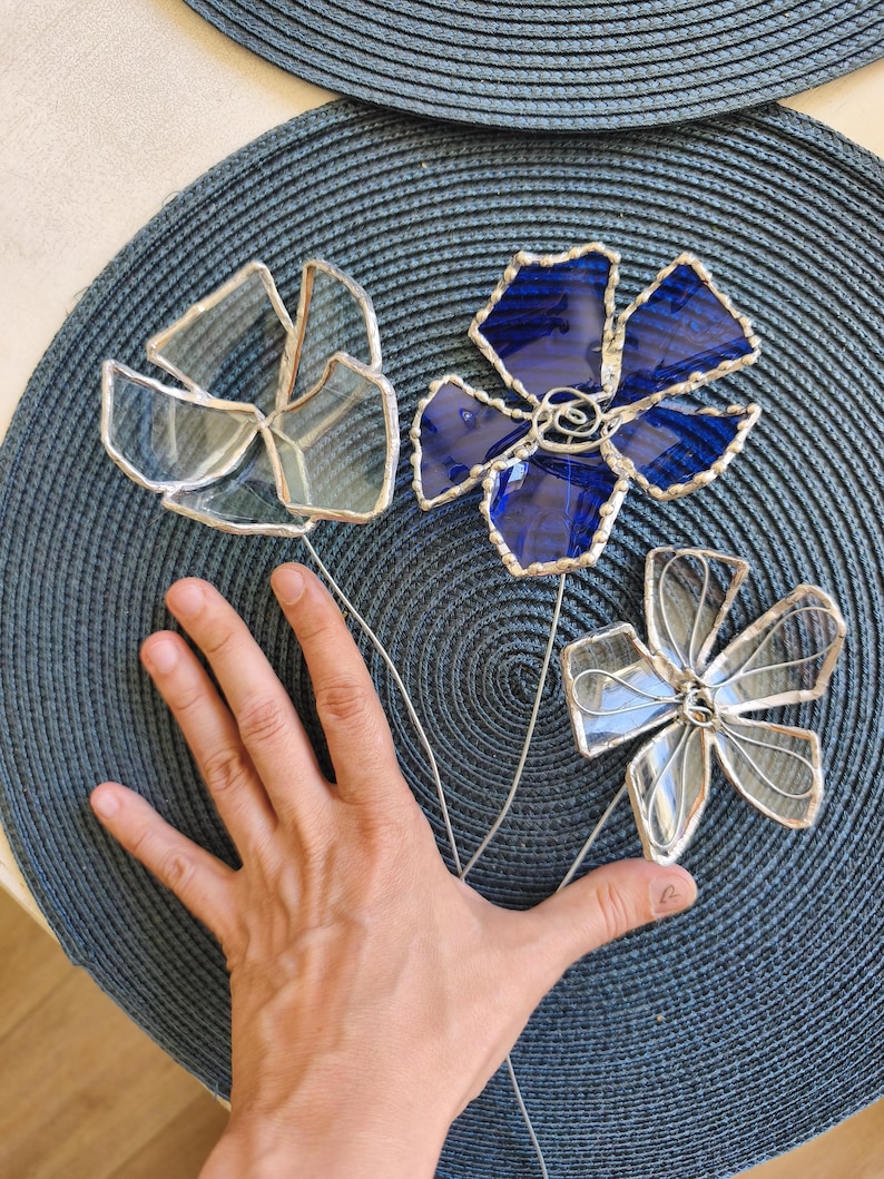 May include: Three stained glass flower sculptures. One is clear, one is blue, and one is clear with silver solder. Each flower has a wire stem. The flowers are arranged on a blue textured surface.