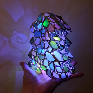 K&ouml;nnte beinhalten: Eine kleine, beleuchtete Glasskulptur in Form eines Baumes. Die Skulptur besteht aus vielen kleinen, bunten Glassplittern, darunter Gr&uuml;n, Blau und Lila. Die Skulptur wird von innen beleuchtet und erzeugt ein warmes, einladendes Leuchten.