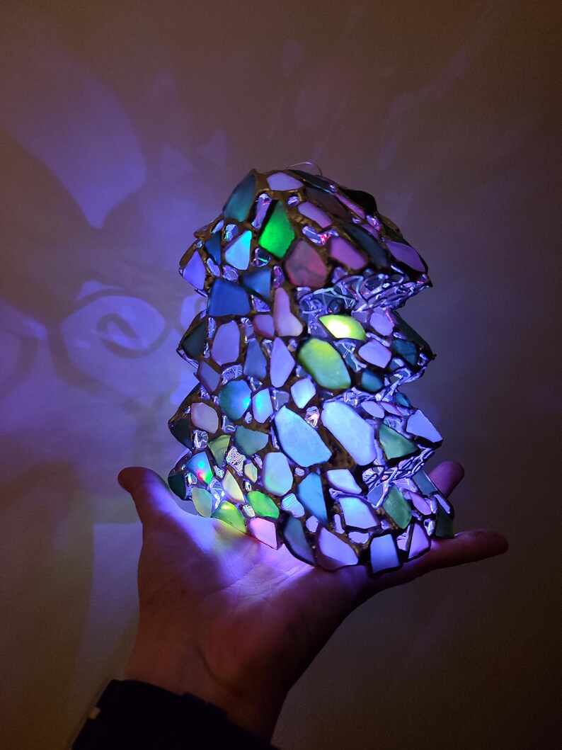K&ouml;nnte beinhalten: Eine kleine, beleuchtete Glasskulptur in Form eines Baumes. Die Skulptur besteht aus vielen kleinen, bunten Glassplittern, darunter Gr&uuml;n, Blau und Lila. Die Skulptur wird von innen beleuchtet und erzeugt ein warmes, einladendes Leuchten.