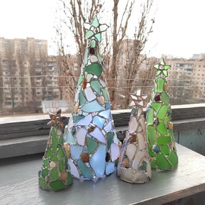 Pode incluir: Uma cole&ccedil;&atilde;o de cinco &aacute;rvores de Natal decorativas. Cada &aacute;rvore &eacute; feita de vitral em tons de verde, azul e branco, com estrelas no topo. As &aacute;rvores variam em tamanho e est&atilde;o dispostas sobre uma superf&iacute;cie cinza.