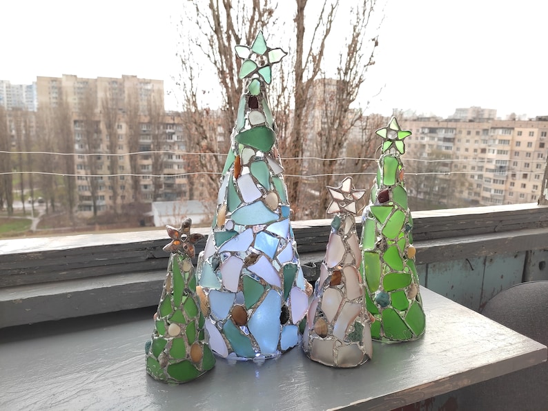 Pode incluir: Uma cole&ccedil;&atilde;o de cinco &aacute;rvores de Natal decorativas. Cada &aacute;rvore &eacute; feita de vitral em tons de verde, azul e branco, com estrelas no topo. As &aacute;rvores variam em tamanho e est&atilde;o dispostas sobre uma superf&iacute;cie cinza.