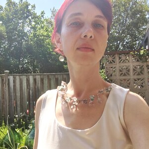May include: A woman wearing a delicate necklace with small, translucent, and colorful beads. The necklace is silver-toned and appears to be handmade. The woman is wearing a cream-colored top, and the background includes a wooden fence and greenery.
