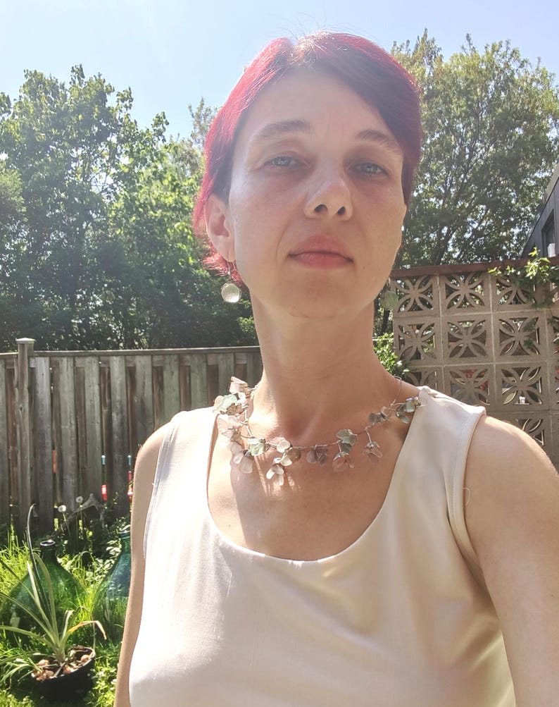 May include: A woman wearing a delicate necklace with small, translucent, and colorful beads. The necklace is silver-toned and appears to be handmade. The woman is wearing a cream-colored top, and the background includes a wooden fence and greenery.