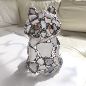 May include: A handcrafted cat figurine made of fused glass pieces in shades of white, grey, and clear. The cat has orange eyes and is outlined with a silver metal. The figurine is sitting on a white surface.