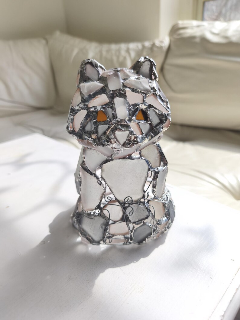 May include: A handcrafted cat figurine made of fused glass pieces in shades of white, grey, and clear. The cat has orange eyes and is outlined with a silver metal. The figurine is sitting on a white surface.