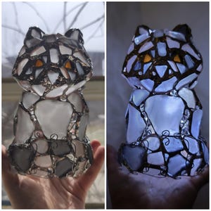 May include: A mosaic cat figurine made from grey, white, and clear glass pieces. The cat's eyes are orange. The figurine is lit from within, creating a soft glow. The cat is approximately 15 cm tall.