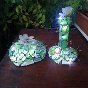 May include: Two green glass mosaic cactus sculptures with white flowers on top. The cactus on the right is tall and slender, while the cactus on the left is short and round. Both cactus sculptures are illuminated from within.