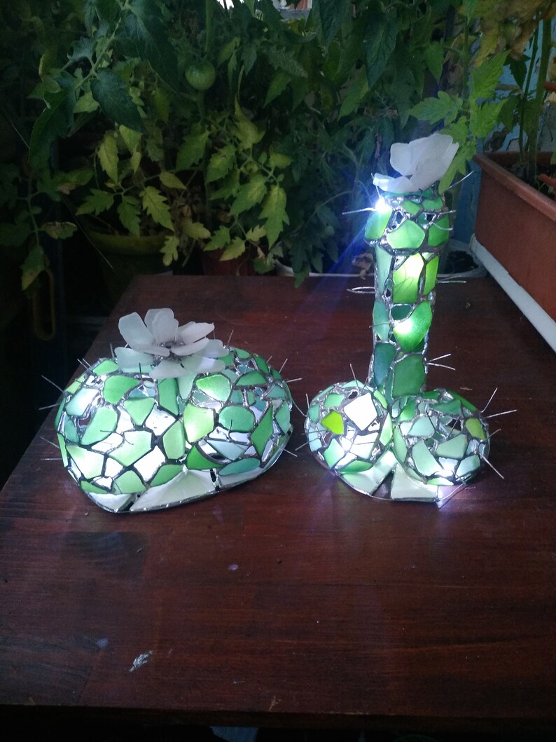 May include: Two green glass mosaic cactus sculptures with white flowers on top. The cactus on the right is tall and slender, while the cactus on the left is short and round. Both cactus sculptures are illuminated from within.