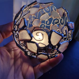May include: A white and silver candle holder made of glass and metal. The holder is shaped like a sphere and has a small candle inside. The candle is lit and casting a warm glow.