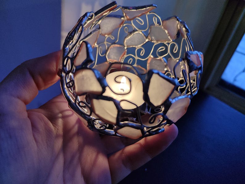 May include: A white and silver candle holder made of glass and metal. The holder is shaped like a sphere and has a small candle inside. The candle is lit and casting a warm glow.