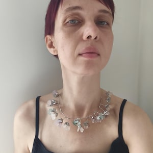 May include: A delicate necklace featuring a silver wire design adorned with small, translucent, flower-shaped beads in shades of pink, green, and clear. The necklace is worn against a black tank top.