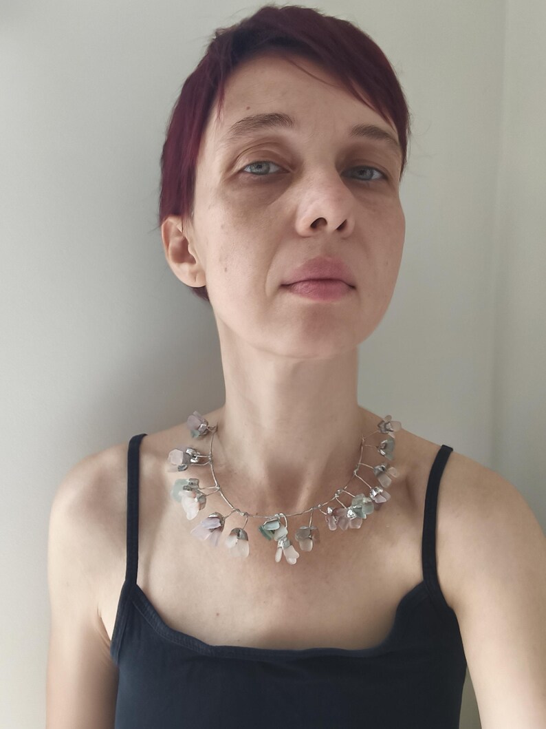 May include: A delicate necklace featuring a silver wire design adorned with small, translucent, flower-shaped beads in shades of pink, green, and clear. The necklace is worn against a black tank top.