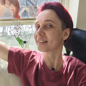 Puede incluir: Una sirena de alambre con la parte superior blanca y la cola verde es sostenida por una persona. La sirena est&aacute; frente a una ventana. La persona lleva una camiseta roja y un collar plateado con un colgante negro.