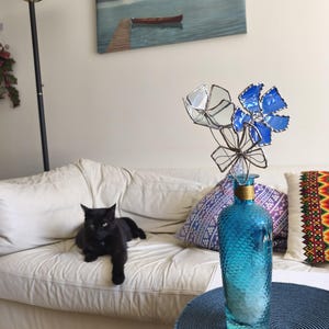 May include: A black cat lounges on a white sofa near a blue glass bottle with decorative flowers. The bottle is on a blue table, and a lake painting hangs on the wall.
