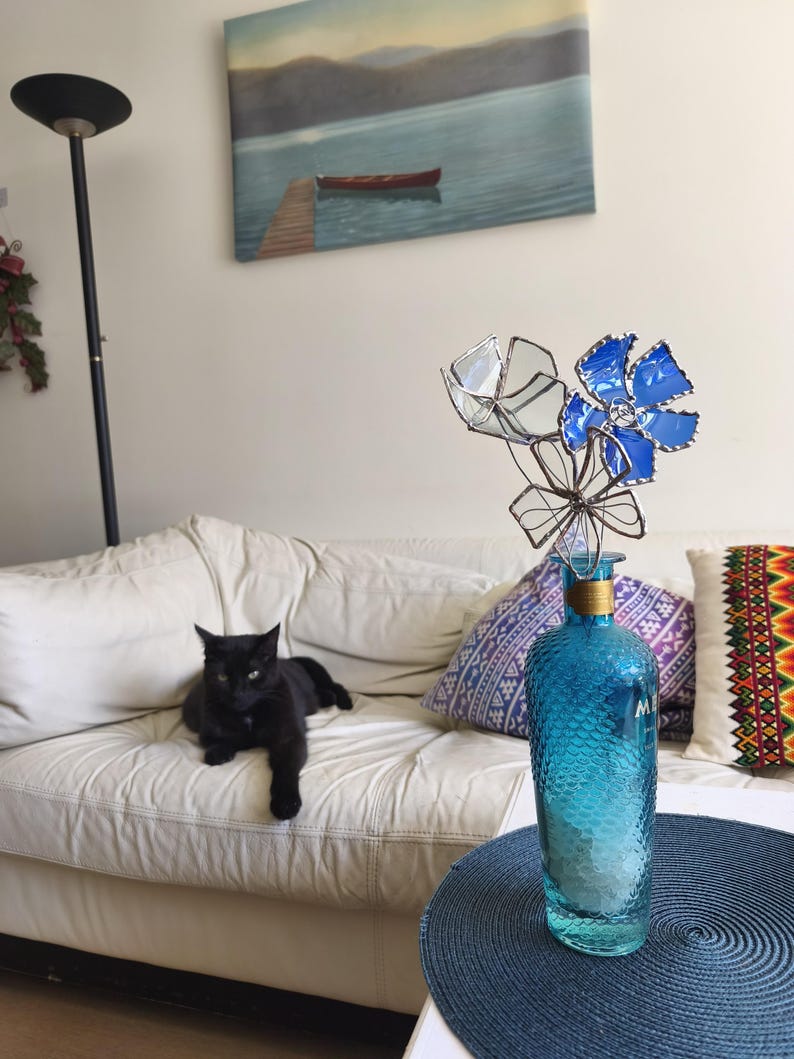 May include: A black cat lounges on a white sofa near a blue glass bottle with decorative flowers. The bottle is on a blue table, and a lake painting hangs on the wall.