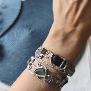 May include: Two silver-toned bracelets with sea glass pieces in shades of brown and green. The bracelets have a decorative, openwork design. The sea glass is set in a silver metal frame, creating a unique and artistic look.