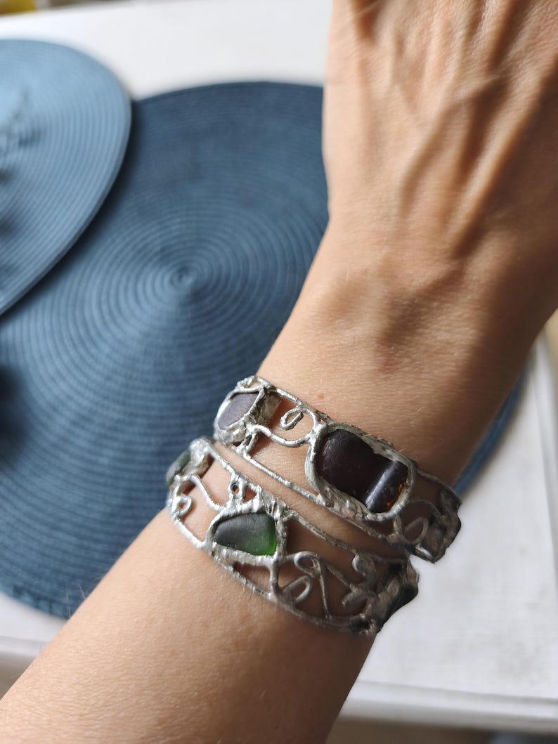 May include: Two silver-toned bracelets with sea glass pieces in shades of brown and green. The bracelets have a decorative, openwork design. The sea glass is set in a silver metal frame, creating a unique and artistic look.