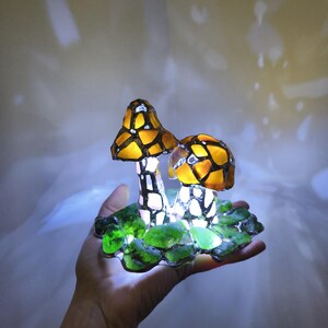 May include: A decorative mushroom lamp with two mushroom caps in shades of yellow and orange, with a mosaic-like pattern. The stems are white and black. The base is green, and the lamp is lit, creating a soft glow.