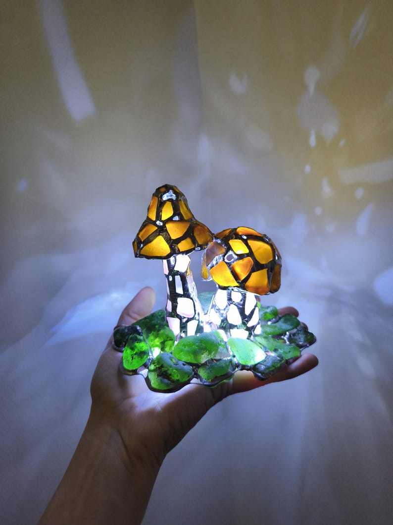 May include: A decorative mushroom lamp with two mushroom caps in shades of yellow and orange, with a mosaic-like pattern. The stems are white and black. The base is green, and the lamp is lit, creating a soft glow.