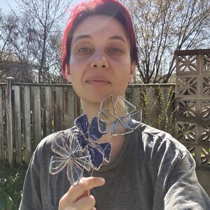 May include: A handcrafted stained glass flower with a blue and clear petal design. The flower is held up by a wooden stem. The glass flower is a decorative item.