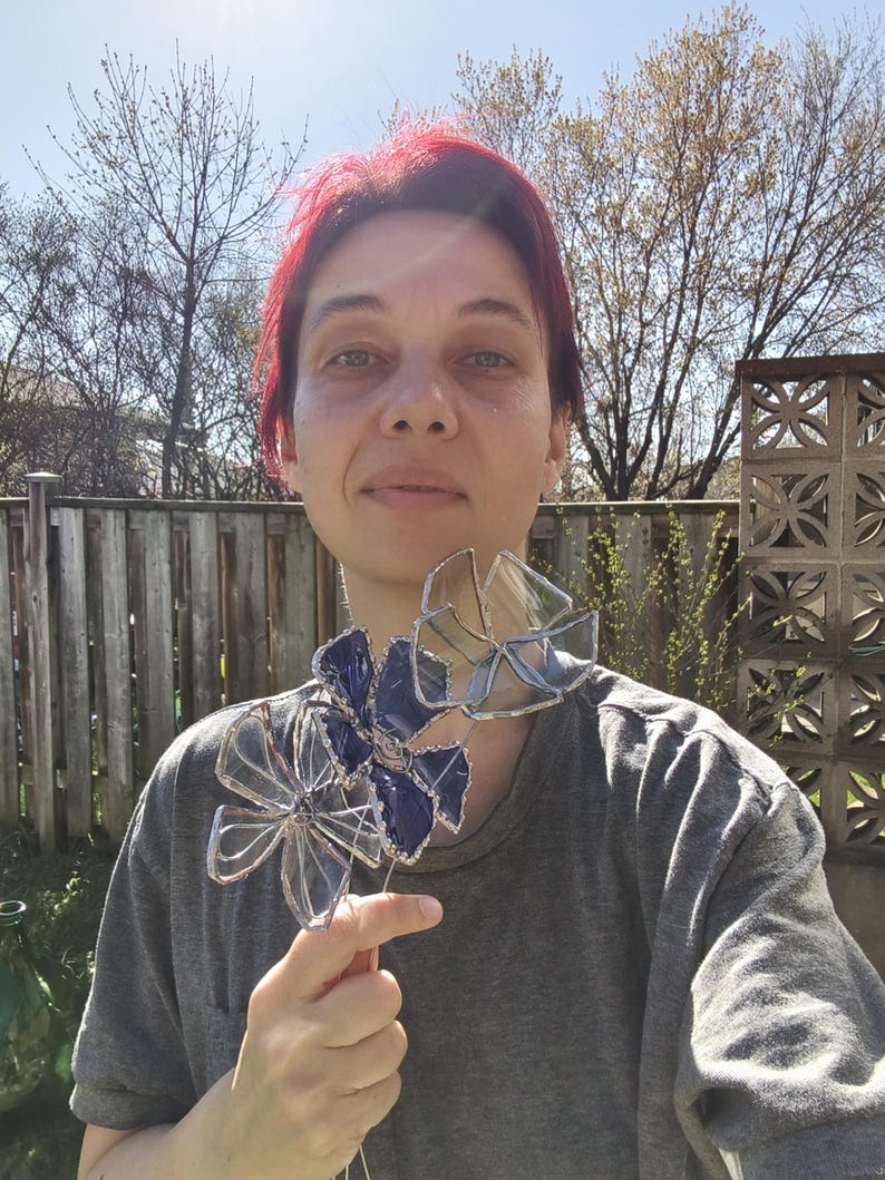 May include: A handcrafted stained glass flower with a blue and clear petal design. The flower is held up by a wooden stem. The glass flower is a decorative item.