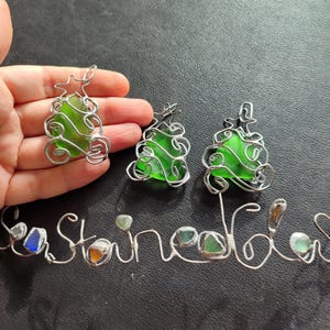 May include: Three green glass Christmas tree ornaments wrapped in silver wire. The ornaments are hanging from a silver wire that spells out the word "Sea Glass".
